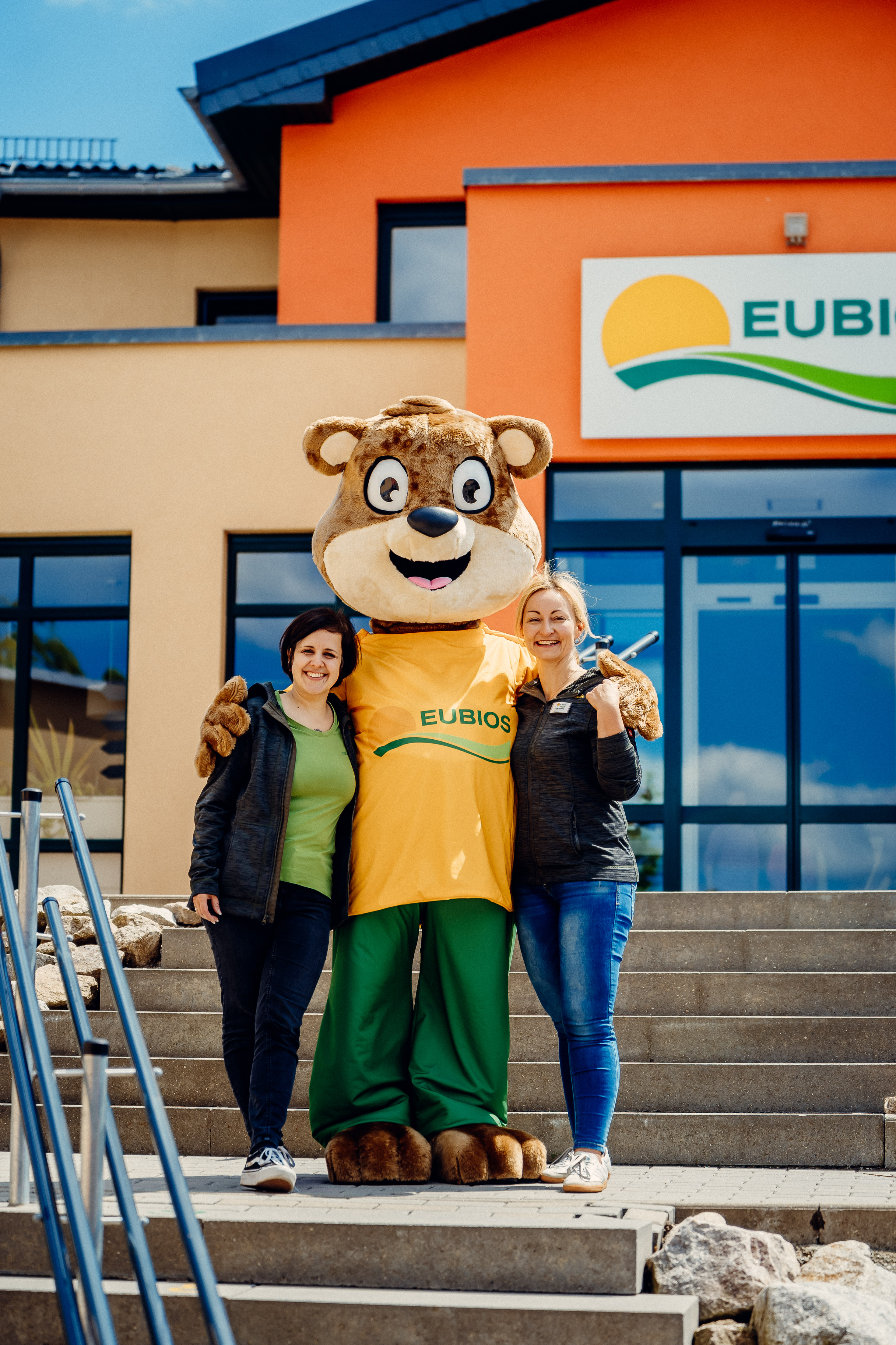 Zwei Mitarbeiterinnen begrüßen gemeinsam mit dem EUBIOS-Maskottchen vor dem Klinikgebäude